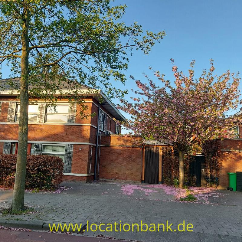 Zweigeschossiges Backsteingebäude mit geschlossenen Holztoren, Bäumen und rosa Blüten am Gehweg.