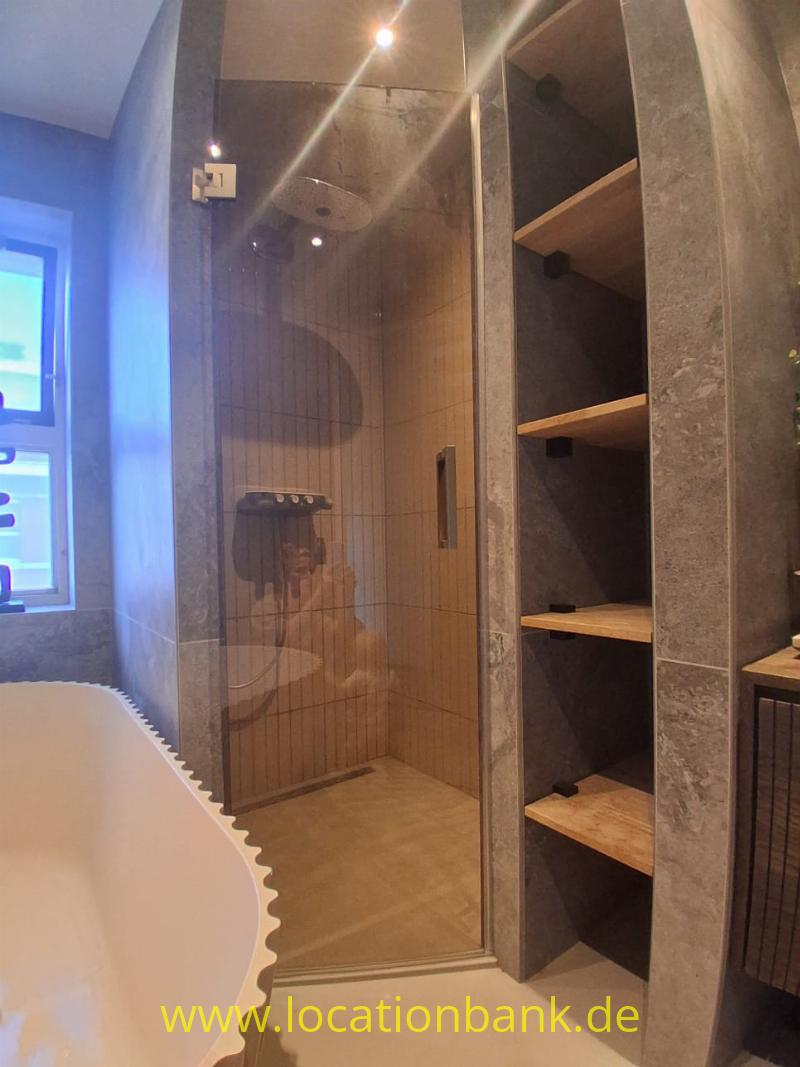 Begehbare Dusche mit Glastür, holzähnlicher Wandverkleidung und offenen Holzregalen neben einer freistehenden Badewanne.