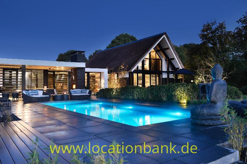 Reetgedecktes Haus mit verglaster Giebelfassade, beleuchtetem Swimmingpool, Terrasse und Skulptur in der Dämmerung.