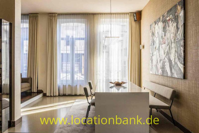Esszimmer mit einem weißen Tisch, Stühlen, großen Fenstern mit Vorhängen und einem Wandkunstwerk.