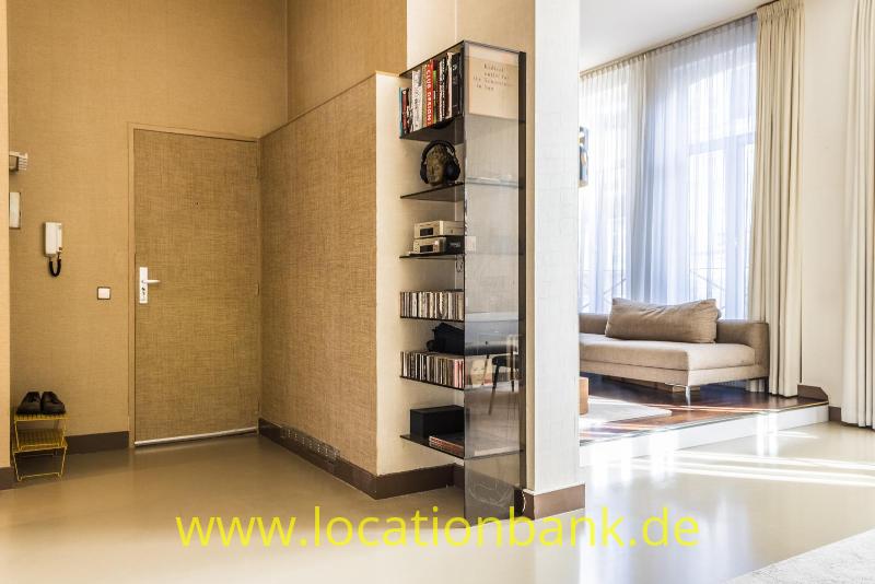 Ein Flur mit einem gläsernen Bücherregal bietet durch einen offenen Durchgang den Blick auf ein Wohnzimmer mit hohen Fenstern.