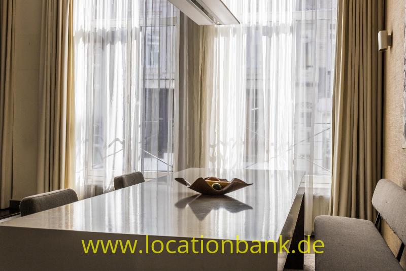 Langer Tisch mit Stühlen, einer Schale und hohen Fenstern mit transparenten Vorhängen.