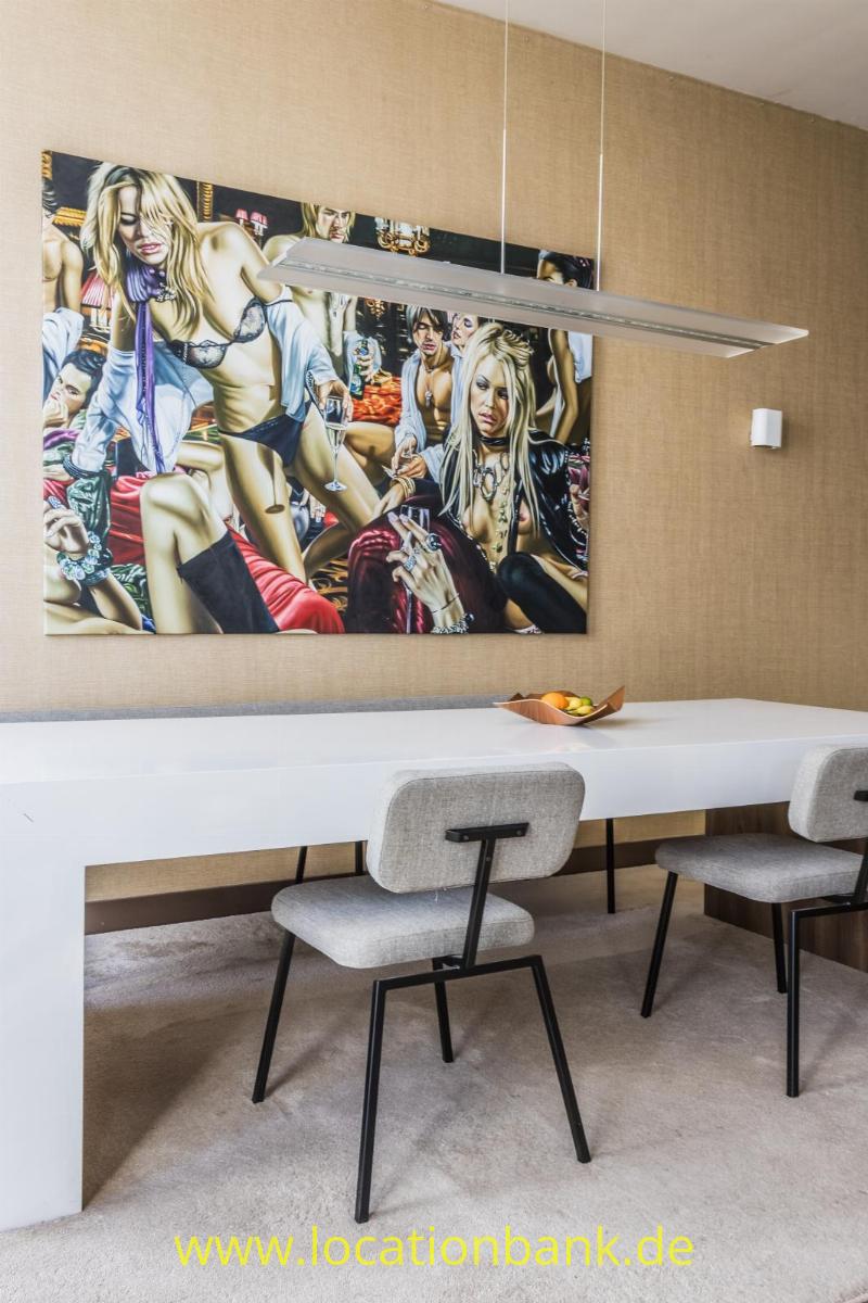 Bunte Wandkunst hängt über einem langen weißen Tisch mit Stühlen und einer länglichen Pendelleuchte.