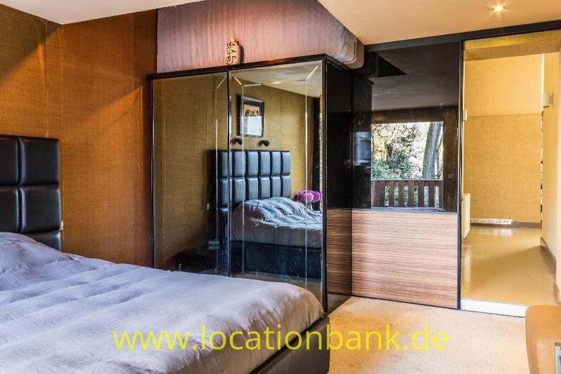 Schlafzimmer mit einem großen Bett, einem verglasten Kleiderschrank und einem offenen Durchgang zu einem Balkon.