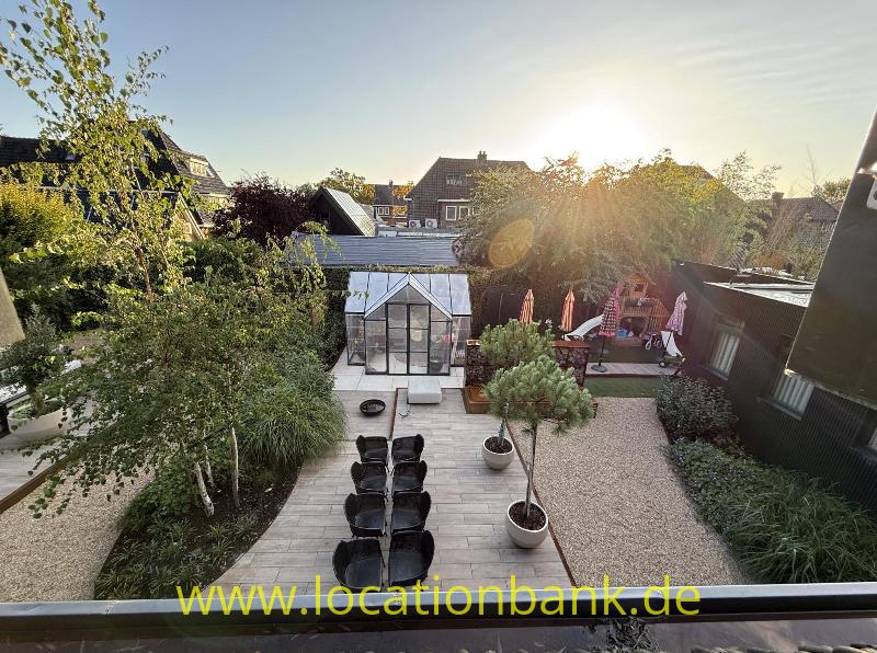 Eine Terrasse zeigt ein gläsernes Gewächshaus, einen langen Tisch mit Stühlen, Pflanzgefäße und eine Kiesfläche.