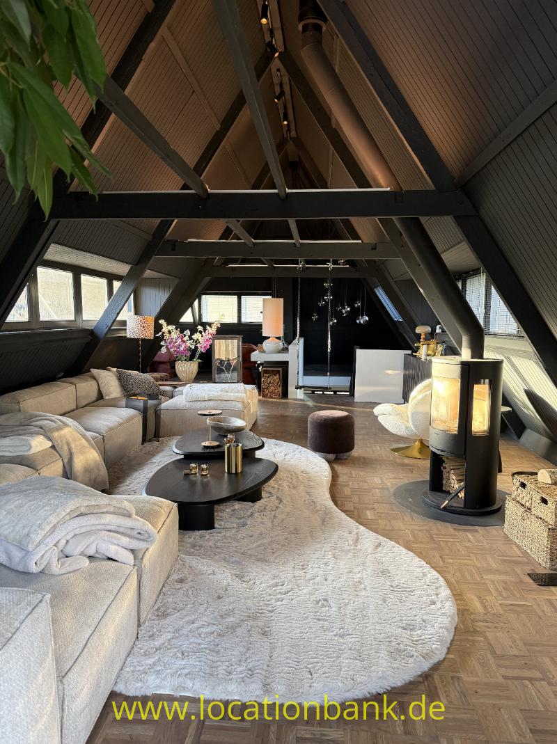 Helles Wohnzimmer unter einem schrägen Dach mit sichtbaren Holzbalken, Sitzbereich und großem Teppich.