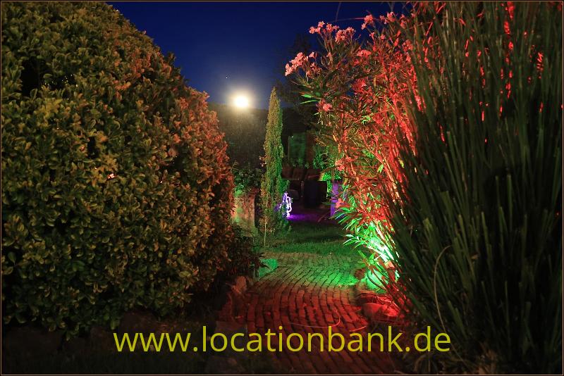 Ein beleuchteter Gartenweg aus Backsteinen verläuft zwischen hohen Hecken und Ziergräsern unter farbigem Abendlicht.
