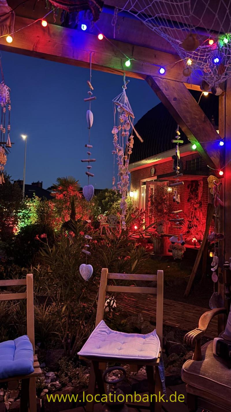 Eine Holzterrasse mit bunten Lichterketten, hängenden Dekorationen, Pflanzen und mehreren Stühlen in der Dämmerung.