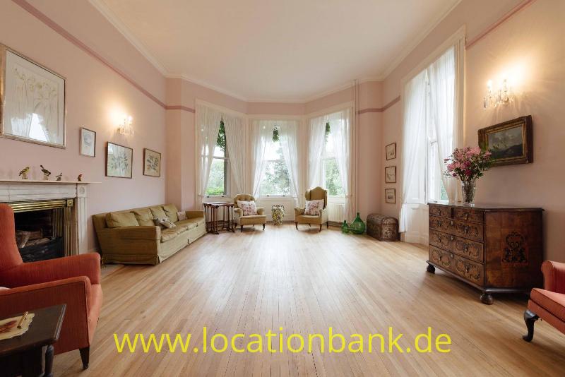 Geräumiges Zimmer mit einem Erker, Holzboden, Kamin, Sitzmöbeln und einer verzierten Kommode.