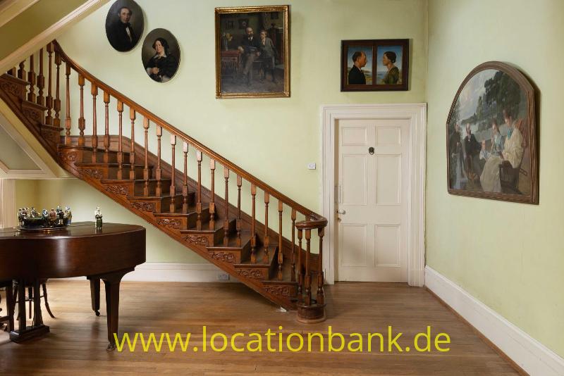 Holztreppe mit detaillierter Balustrade neben einem Flügel, Gemälden und Porträts in einem Innenraum.