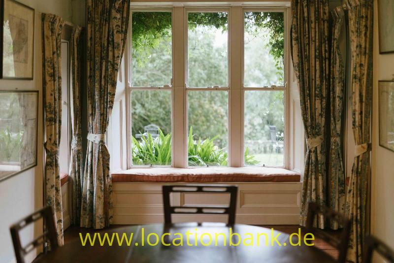 Ein großes Holzfenster mit vier Scheiben, eingerahmt von gemusterten Vorhängen, bietet den Blick auf einen grünen Garten.