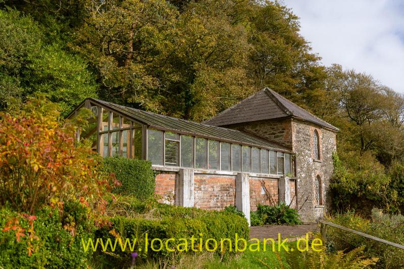 Ein langer Glasanbau mit Backsteinsockel grenzt an ein Steingebäude mit Schieferdach und ist von Bäumen und Sträuchern umgeben.