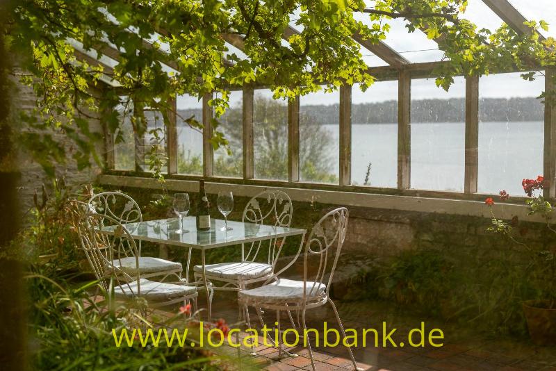 Überdachte Terrasse in Gewächshaus mit Glaswänden, einem Metalltisch und Stühlen, umgeben von Grün am Wasser.