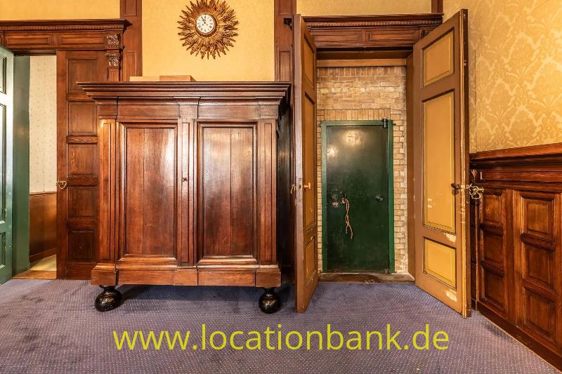 Ein hoher Holzschrank steht unter einer Wanduhr, neben einer geöffneten Holztür und einer grünen Tür in einer Backsteinwand.