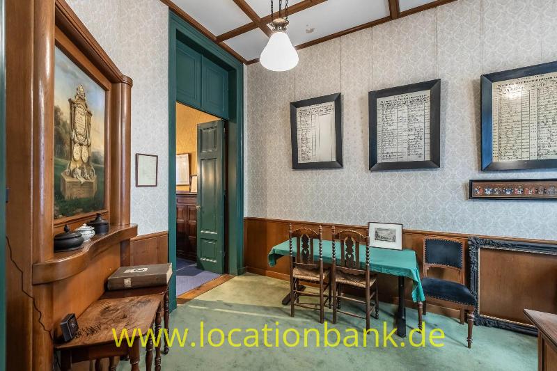 Innenraum mit einer hölzernen Vitrine, einer grünen Tür, gerahmten Dokumenten und einem Tisch mit Stühlen.