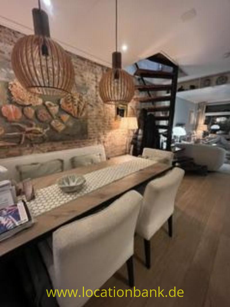 Essbereich mit einem langen Holztisch, gepolsterten Stühlen, Pendelleuchten und einem Wandgemälde neben einer offenen Treppe.