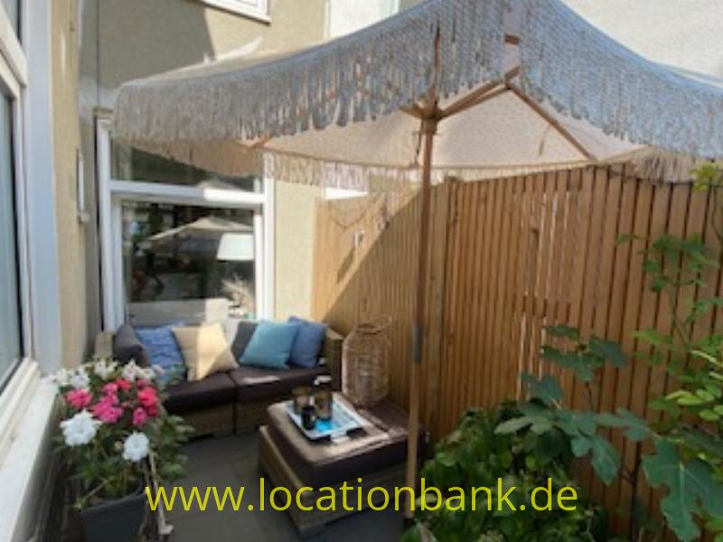 Eine Sitzecke auf einer Terrasse unter einem Sonnenschirm mit Fransen, mit einem Holzzaun und blühenden Pflanzen.