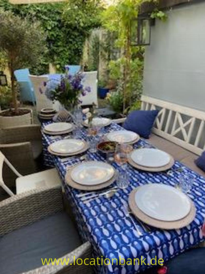 Gedeckter Tisch im Freien mit blau gemusterter Tischdecke, Geschirr, Gläsern und Blumen, umgeben von Stühlen, Pflanzen und einer weißen Wand.