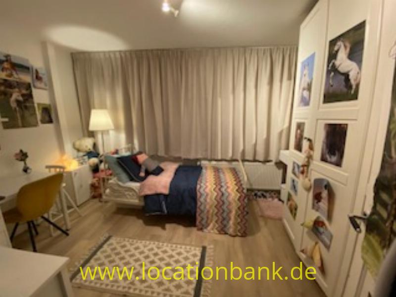 Schlafzimmer mit einem bezogenen Bett, geschlossenen Vorhängen, einem Schreibtisch und gerahmten Fotografien an den Wänden.