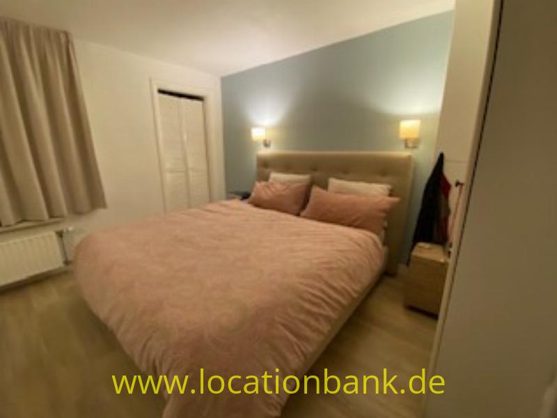 Ein gemachtes Doppelbett mit einer rosafarbenen Tagesdecke steht an einer blaugrauen Wand, mit Nachttischen und Wandleuchten.