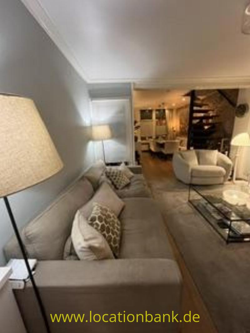Wohnzimmer mit einem beigen Ecksofa, einer Stehlampe, einem Sessel, einem Couchtisch und einer Treppe im Hintergrund.