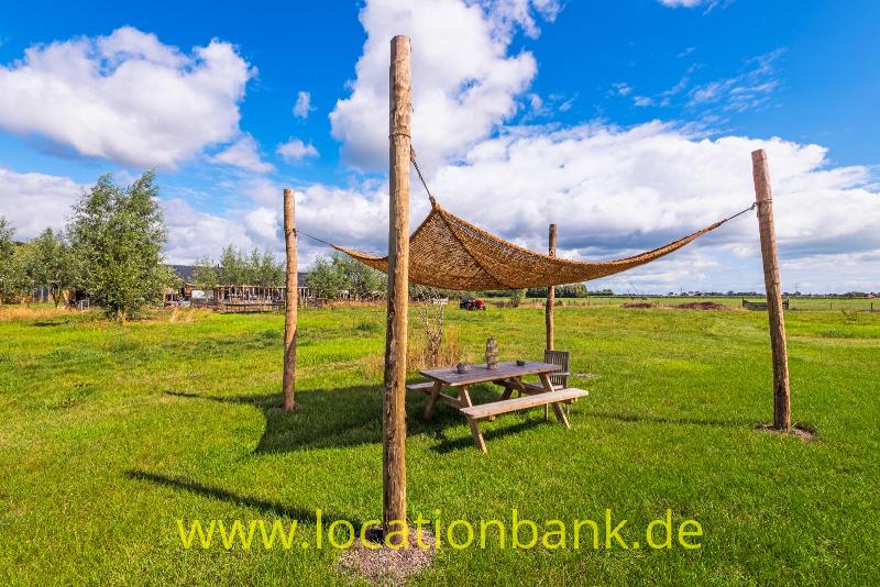 Ein Picknicktisch steht unter einem gespannten Tuch zwischen Holzpfosten auf einer offenen Wiese.