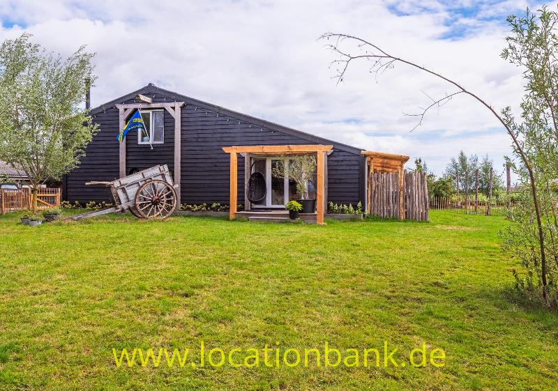Dunkles Holzgebäude mit überdachter Veranda, Holzkarren und weitläufigem grünen Garten unter einem bewölkten Himmel.
