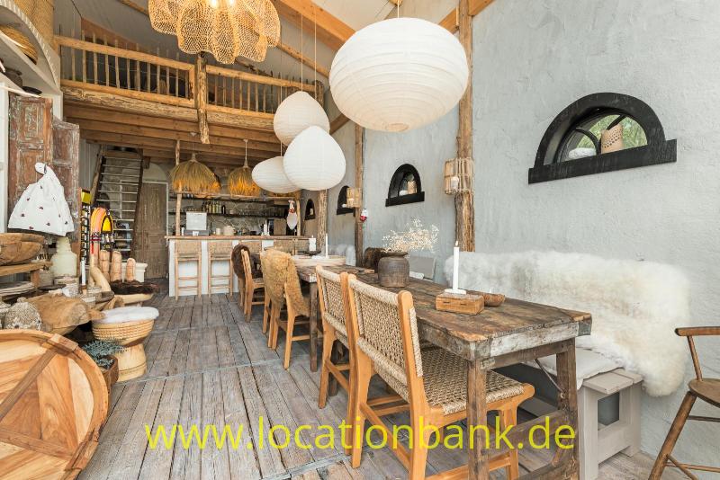 Langer Holztisch mit geflochtenen Stühlen, Hängelampen und kleinen Bogenfenstern an einer blaugrauen Wand.