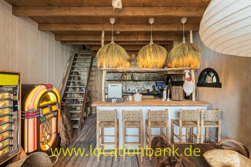 Holztheke mit hohen Hockern, drei geflochtenen Hängeleuchten und einer bunten Jukebox unter Holzbalken.