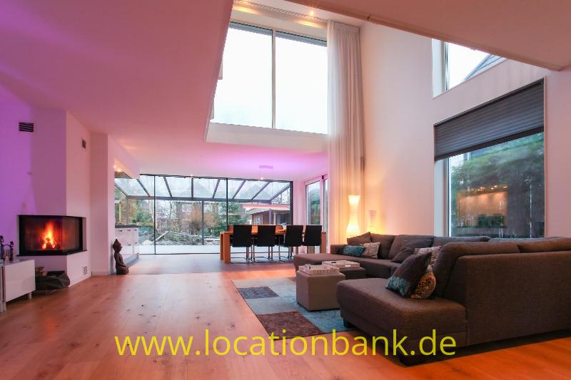Geräumiges Wohnzimmer mit Ecksofa, Kamin, Essbereich und großen Fenstern mit Blick ins Grüne.