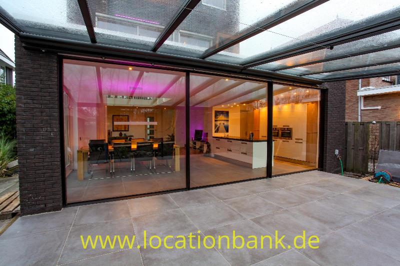 Verglaste Erweiterung mit transparentem Dach, sichtbarer Küche und Essecke, angrenzend an eine gepflasterte Terrasse.