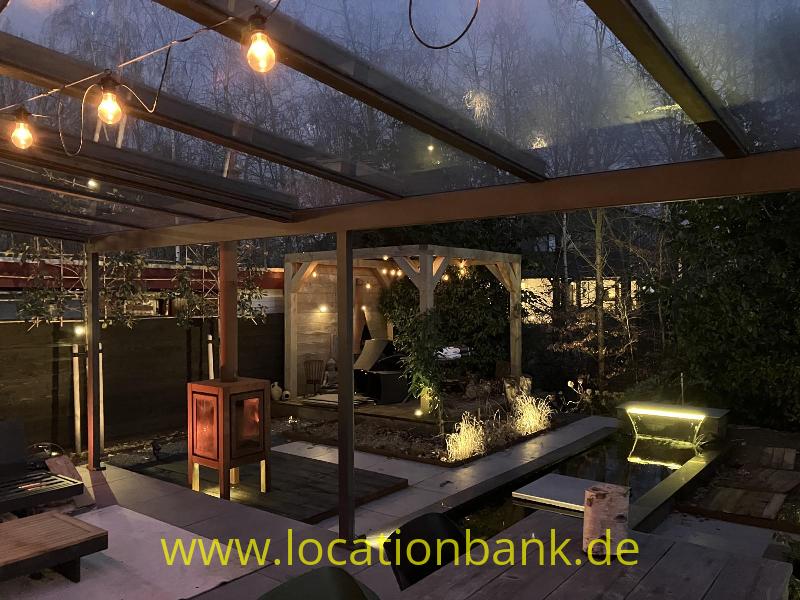 Überdachte Terrasse mit Lichterketten, einem Teich und beleuchteten Pflanzen in einem Garten.