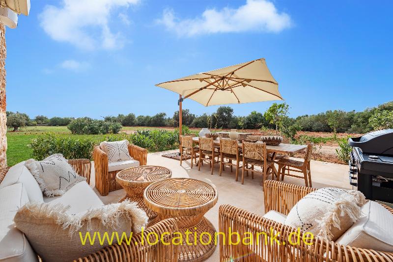 Terrasse mit Rattansitzmöbeln, Esstisch, Sonnenschirm und Blick auf grüne Felder.