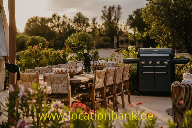 Ein schwarzer Grill steht neben einem langen Holztisch mit geflochtenen Stühlen, umgeben von blühenden Pflanzen.
