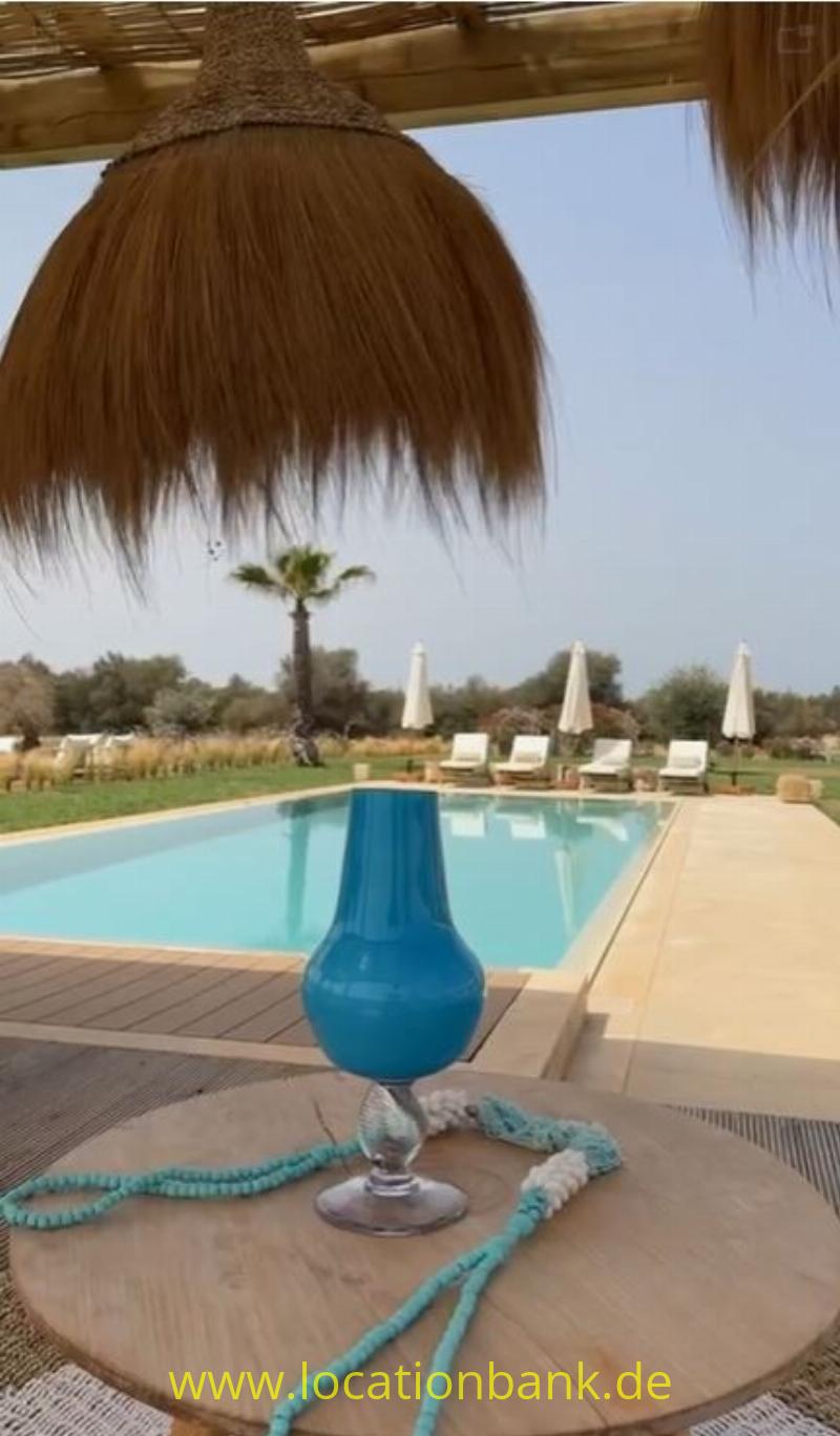 Blaue Vase mit einer türkisen Perlenschnur auf einem runden Holztisch, dahinter ein Swimmingpool und geschlossene Sonnenschirme.