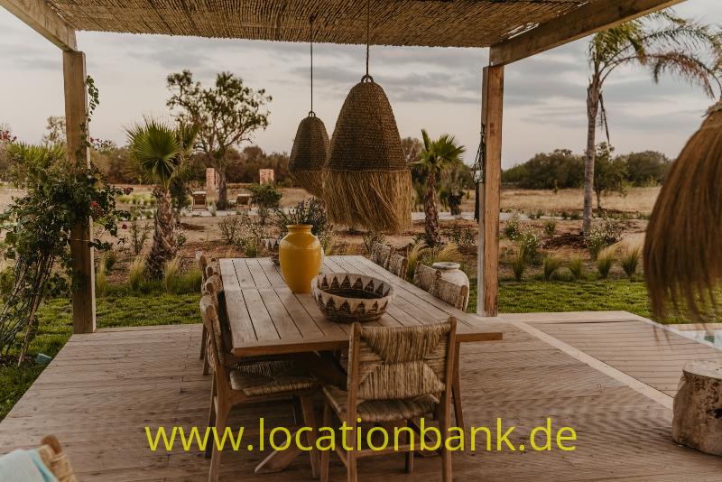 Ein Holz-Esstisch auf einer überdachten Terrasse mit geflochtenen Hängelampen und grüner Landschaft im Hintergrund.
