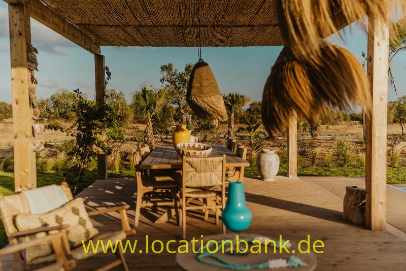 Überdachte Holzterrasse mit Reetdach, Esstisch, türkisfarbener Vase und trockenem Garten im Hintergrund.