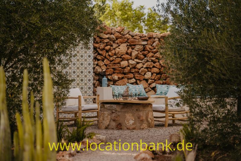 Sitzbereich im Freien mit Holzstühlen, einem niedrigen Steintisch, einer Steinmauer und einer gefliesten Rückwand.