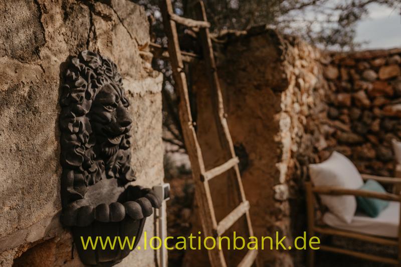 Ein dunkles Löwenkopfrelief hängt an einer verwitterten Steinmauer neben einer Holzleiter.