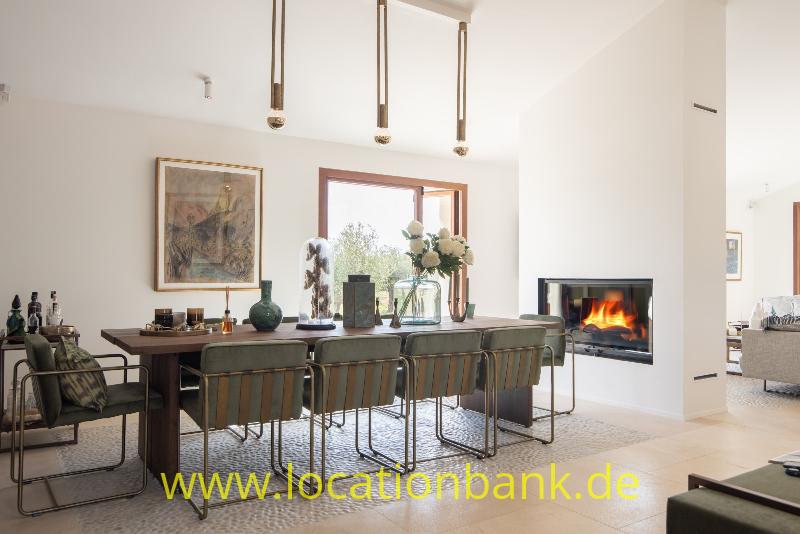 Langer Esstisch mit mehreren Stühlen, drei Pendelleuchten, einem Gemälde und einem brennenden Kamin.
