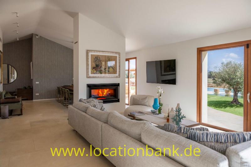 Wohnzimmer mit großem grauem Ecksofa, Kamin, Fernseher und großen Glastüren zu einem Garten mit Olivenbaum.