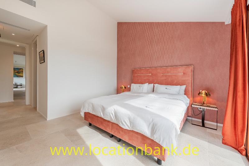 Ein Doppelbett mit weißer Bettwäsche steht vor einer rosaroten Wand neben einem offenen Durchgang.