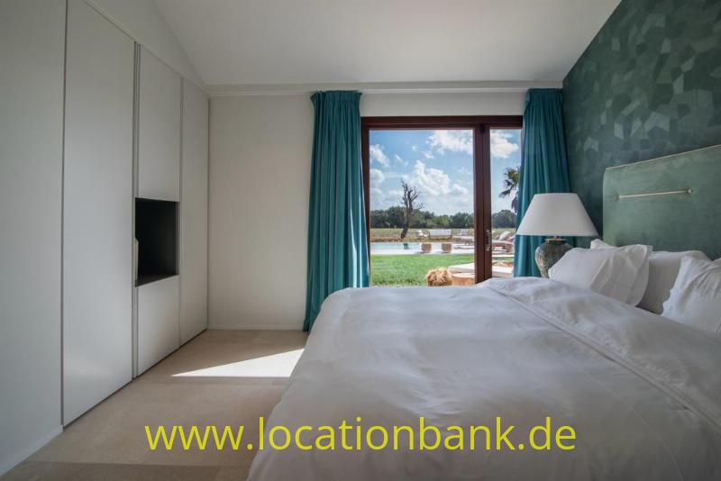 Schlafzimmer mit einem bezogenen Bett, einer grünen gemusterten Wand und einem Fenster mit türkisfarbenen Vorhängen.