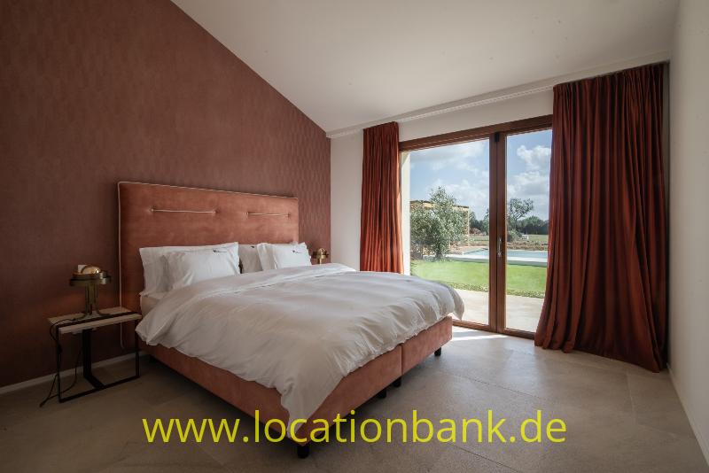 Schlafzimmer mit einem bezogenen Doppelbett, langen Vorhängen und Glastüren zu einem grünen Außenbereich.