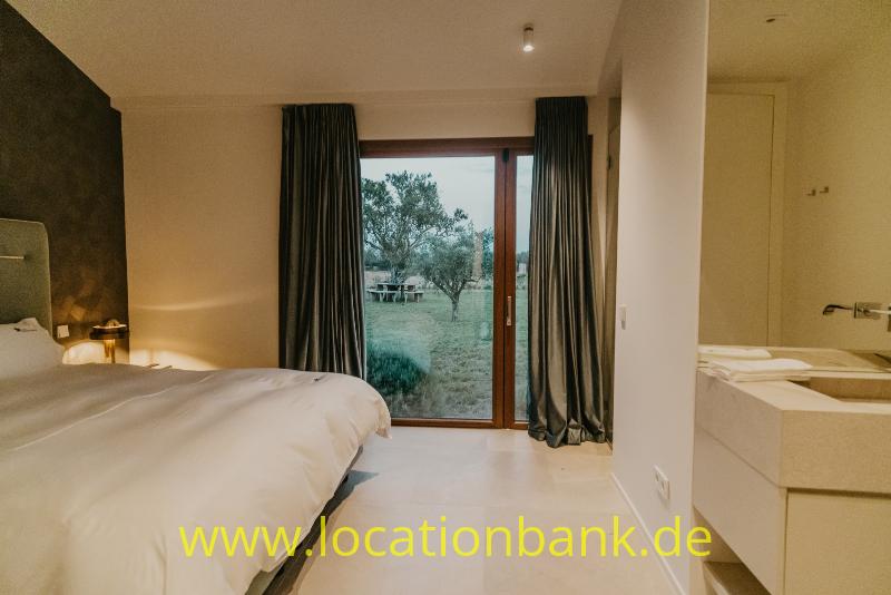 Schlafzimmer mit einem bezogenen Bett, dunklen Vorhängen und einer Glastür mit Blick auf Bäume und Rasen.