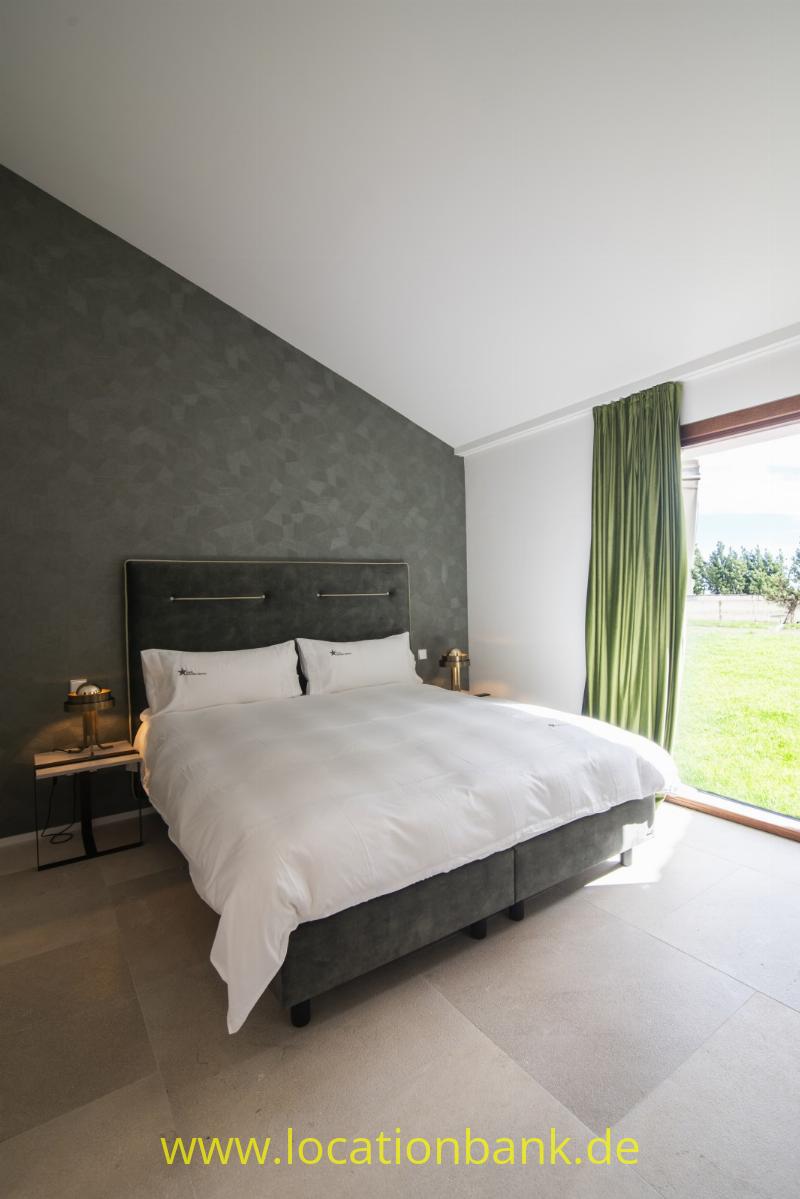 Ein Schlafzimmer mit einem bezogenen Doppelbett vor einer dunklen Strukturtapete und grünen Vorhängen neben einem großen Fenster.
