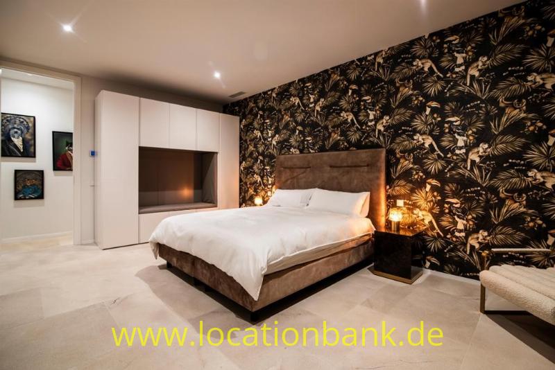 Schlafzimmer mit einem großen Bett, dunkler botanischer Tapete, weißen Schränken und warmer Beleuchtung.