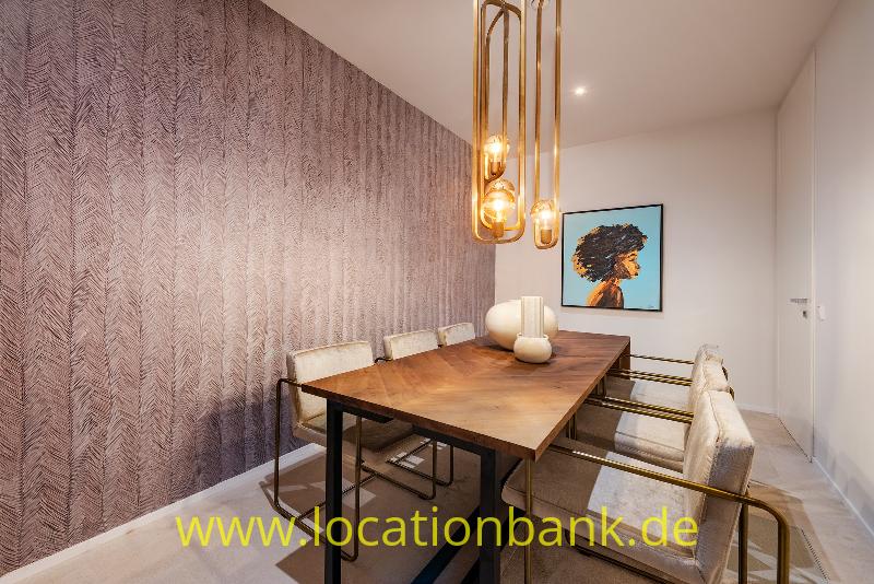 Hölzerner Esstisch mit Stühlen unter gläsernen Pendelleuchten, neben einer Wand mit gemusterter Tapete und einem Porträt.