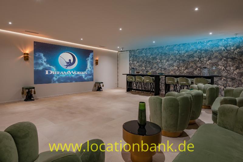Große Leinwand mit einem runden logo in einer Lounge mit grünen Sesseln und einer Bar.