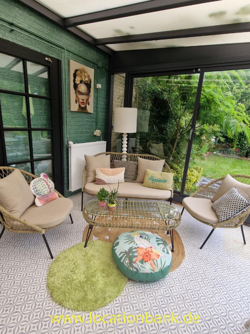 Sitzbereich mit einem beigefarbenen Sofa, zwei Sesseln, einer Glaswand und Blick auf einen grünen Garten.